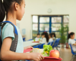 A young girl in a school cafeteria holding a tray of food, looking for a place to sit
