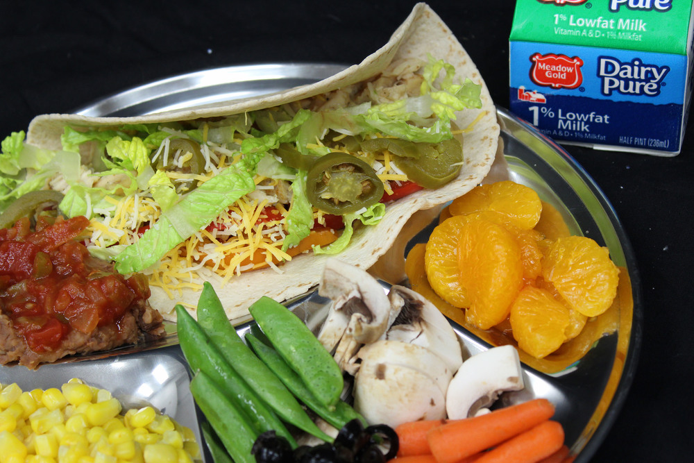 a school lunch of a soft taco with salsa, corn off the cob, tangerine slices, and mixed vegetables