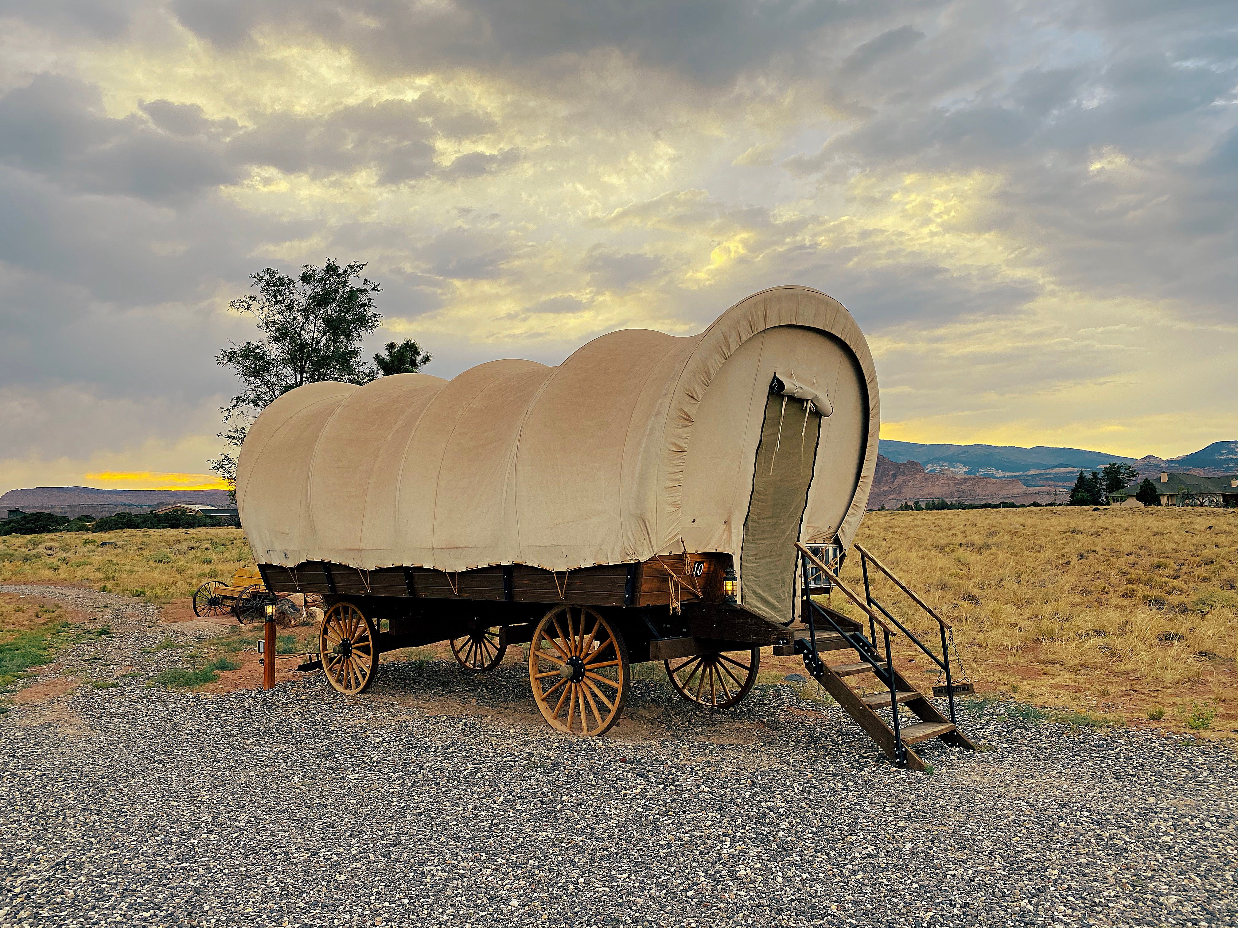 A classic covered wagon sits in a scenic field under a dramatic sunset